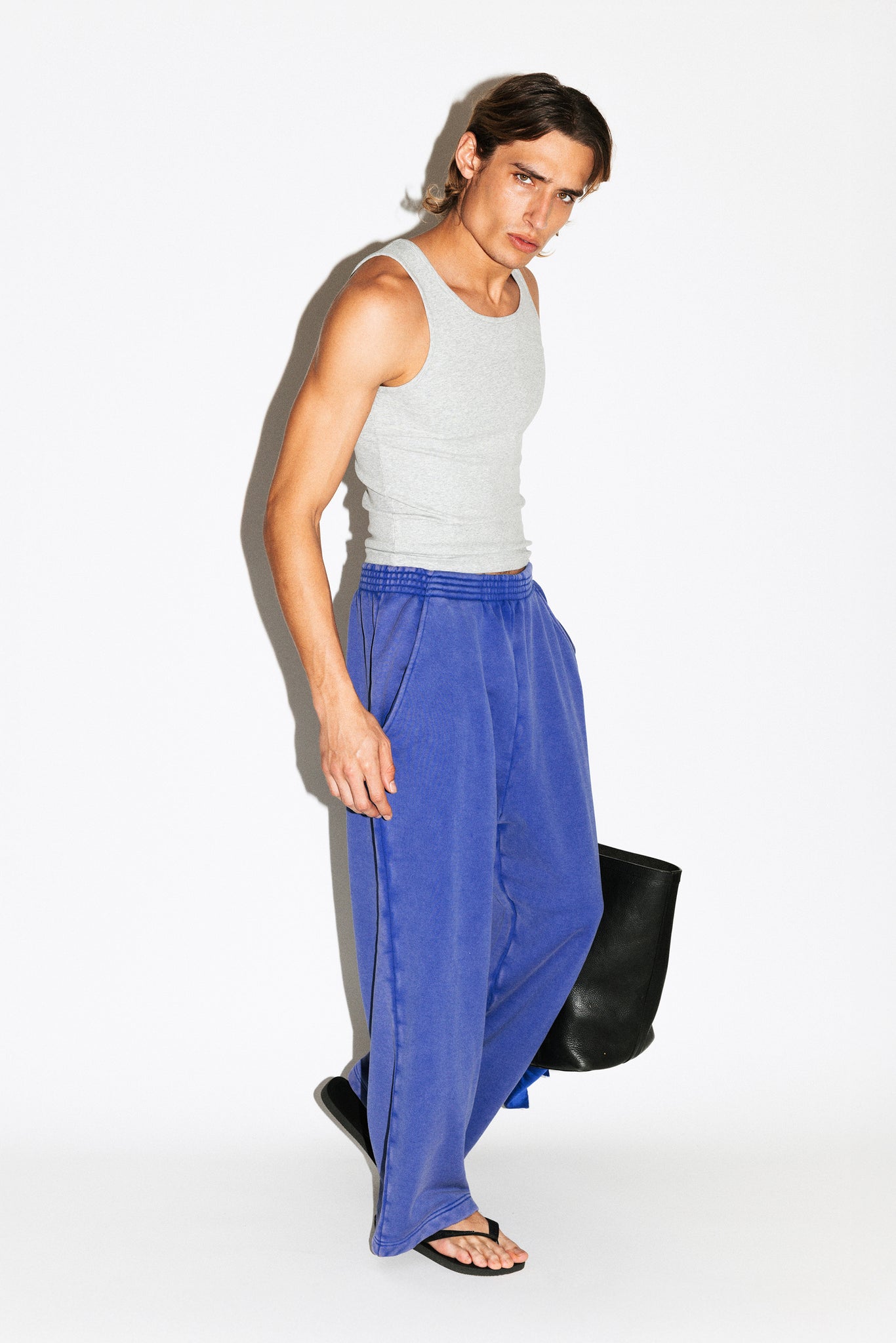Person wearing a gray tank top and blue pants holding a black bag on a white background